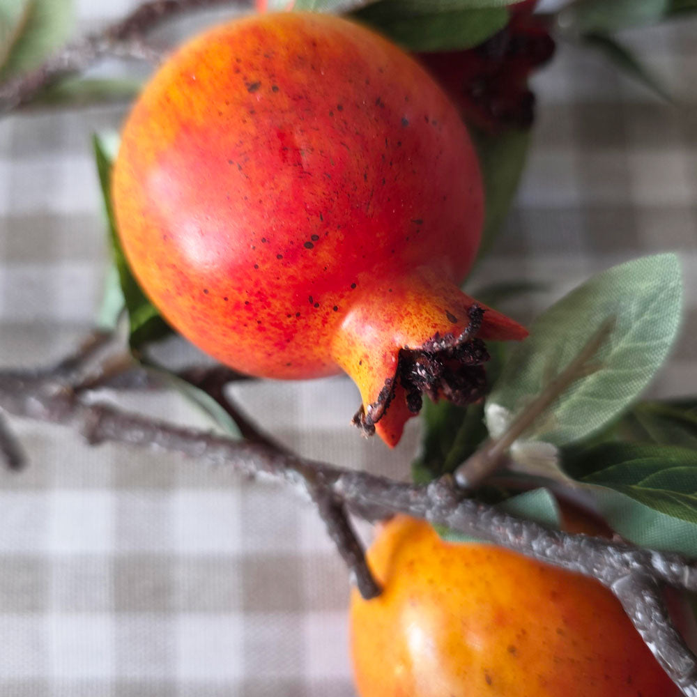 A close-up of a pomegranate on a branch with green leaves, set against a soft beige and white checkered fabric background.