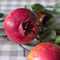 A close-up of a red pomegranate with dark spots and stem scars, resting on a branch with green leaves over a beige-and-white checkered fabric background.