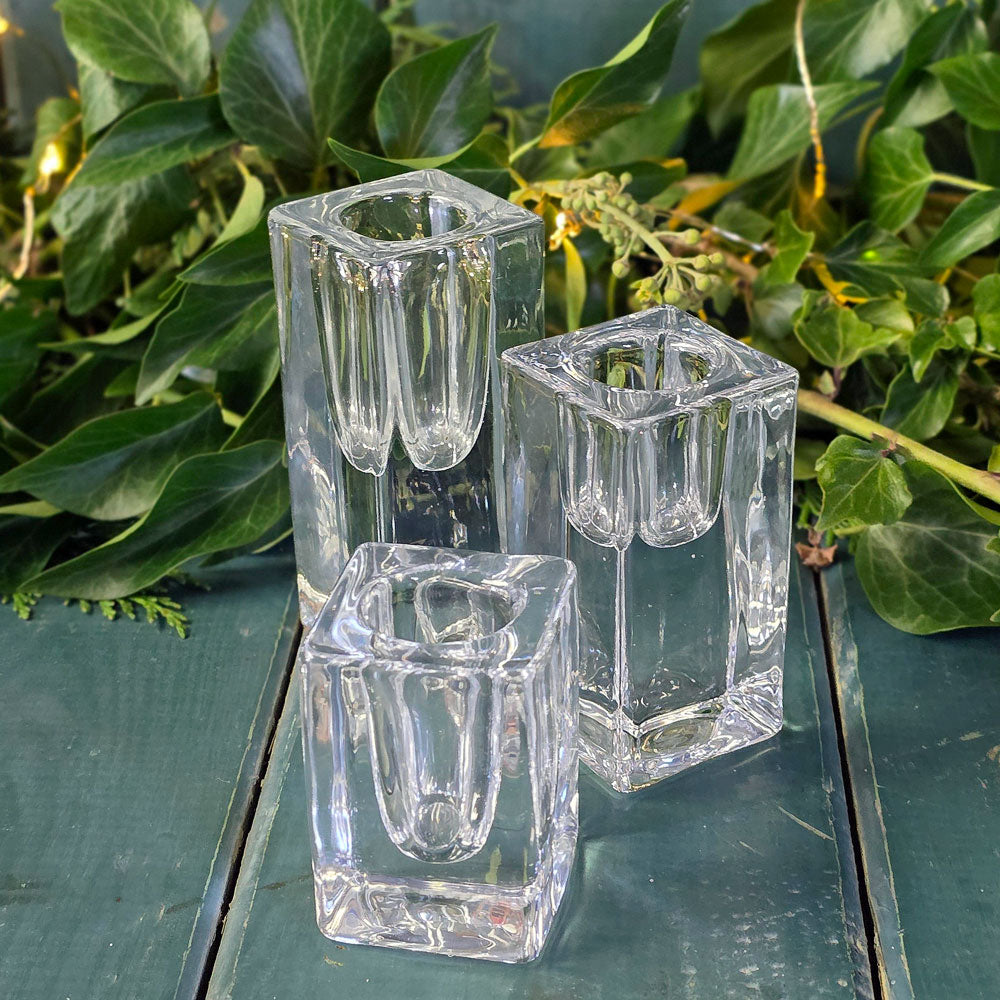 Three clear glass square candle holders on a wooden surface with green foliage in the background