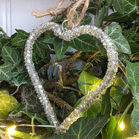 A heart-shaped clear glass hanging ornament wrapped with silver wire and suspended by a rustic twine loop, displayed among green ivy leaves and small warm white string lights.