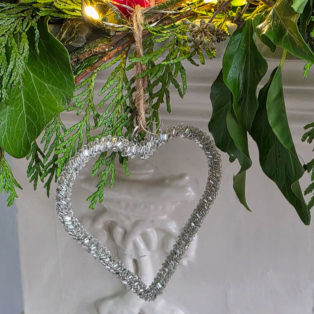Silver glass beaded heart shaped tree decoration, next to greenery.