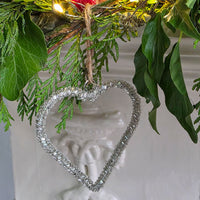 Silver glass beaded heart shaped tree decoration, next to greenery.