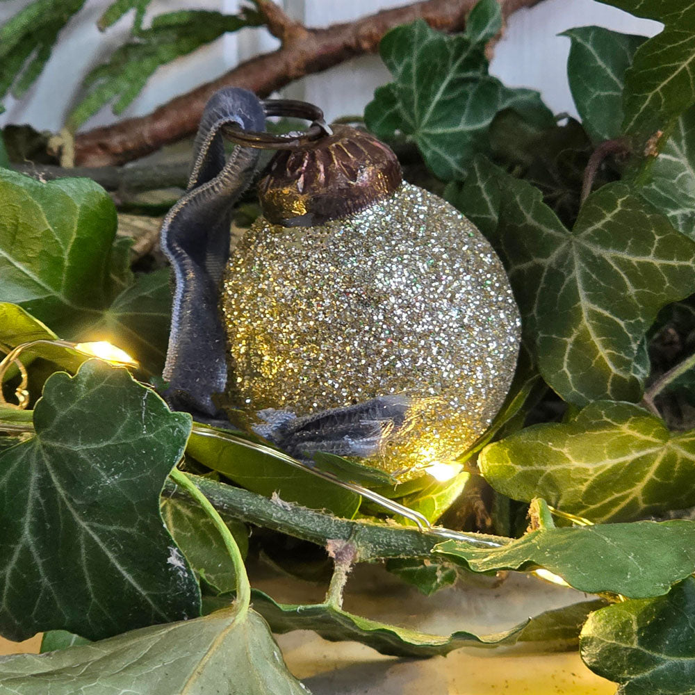 A glittery silver Christmas bauble , nestled among green ivy leaves with warm string lights.