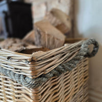 A close-up of a natural wicker basket with a thick gray rope handle, filled with firewood logs, set against a softly blurred rustic background.